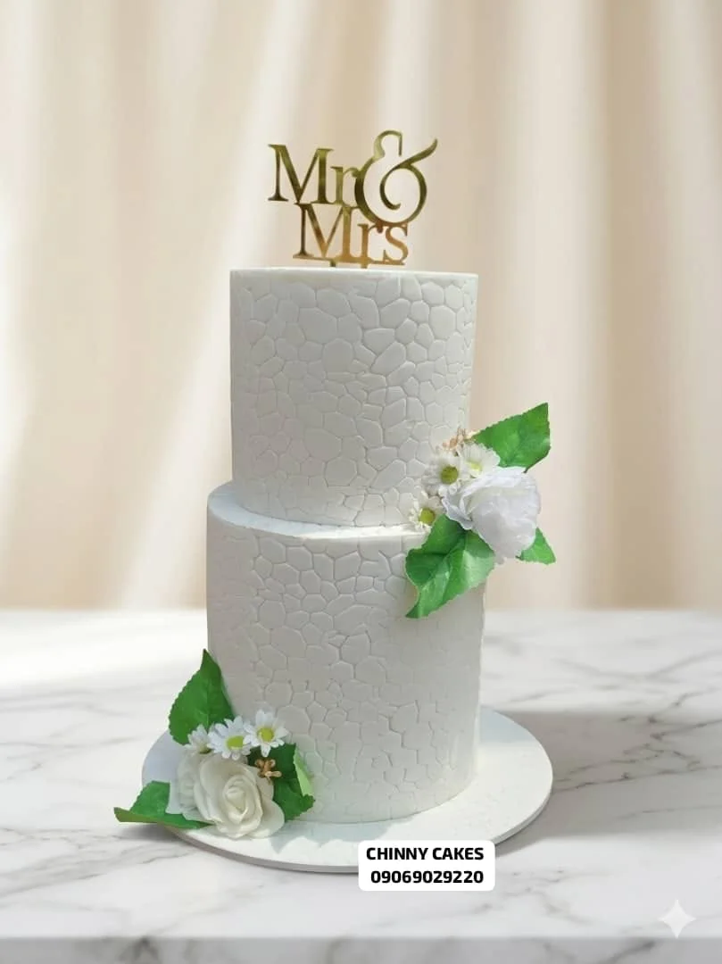 Two-tier white wedding cake with subtle texture, white flowers and green leaves